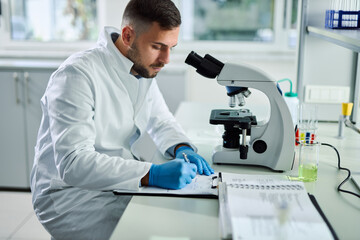 Young male scientist taking notes while working on microscope in laboratory.