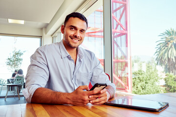 Latin businessman holding smartphone and smiling in modern office, with digital tablet on desk