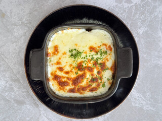 Classic Italian baked casserole with bacon and risotto inside. Topped with melted baked mozzarella cheese. Seasoned with herbs and chives, served on a black plate, grey marble table.