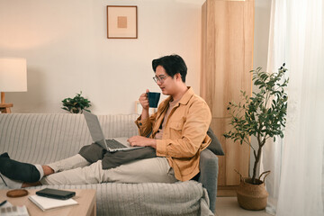 A man sits comfortably on a couch, sipping coffee while working on his laptop in a cozy home, the...