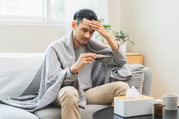 Fototapeta premium Sick, influenza asian young man, male have fever hand holding thermometer, touching forehead for check measure body temperature, illness while sitting rest on sofa at home. Health care on virus person