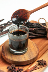 Rich, dark vanilla extract drips from a wooden spoon into a glass jar, surrounded by whole vanilla...