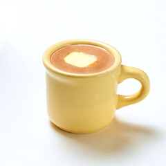 A warm creamy chocolate drink is presented in a bright yellow cup, topped with a slice of butter, against a white backdrop, serving as a comforting treat