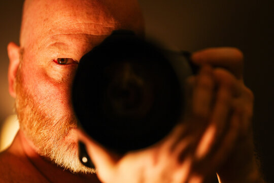 Bald photographer with staring with intensity through his camera to take a photograph at night. The man has a red beard and intense stare while holding the lens. 