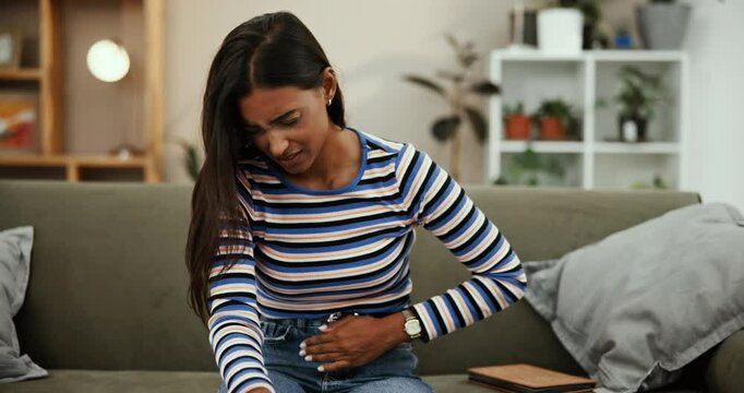 Stomachache, gut and pain for woman, couch and discomfort with bloating in living room and house. Lounge, constipation and uncomfortable with gas on sofa, indigestion and person with cramps in home