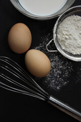 fresh eggs, milk in a plate, flour in a sieve and scattered on the background, a whisk lies on a dark background, different angles
