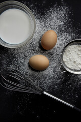 fresh eggs, milk in a plate, flour in a sieve and scattered on the background, a whisk lies on a...