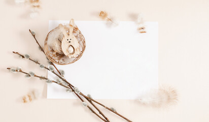 Easter card. Easter holiday composition on beige background with empty white paper sheet for congratulations. Branches of blooming willow, Easter eggs, funny rabbit pendant. Flat lay.