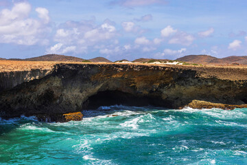 Waves crashing against rocky coastline near natural cave in Arikok National Park under blue sky...