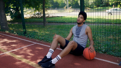 Sweaty African American guy sits on the court, feeling exhausted after a competitive basketball game