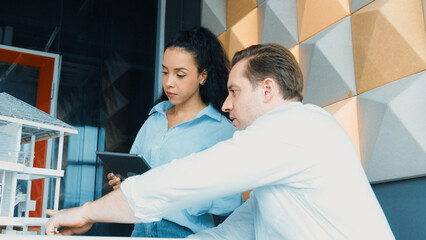Portrait of project manager pointing at house model while discussion about building design. Caucasian cooperative architect engineer team brainstorming about building material. Teamwork. Manipulator.