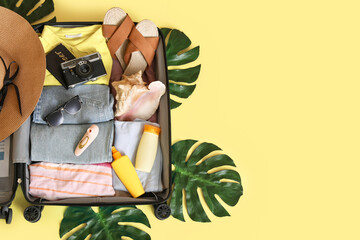 Open suitcase with clothes, beach accessories, vibrator and palm leaves on yellow background