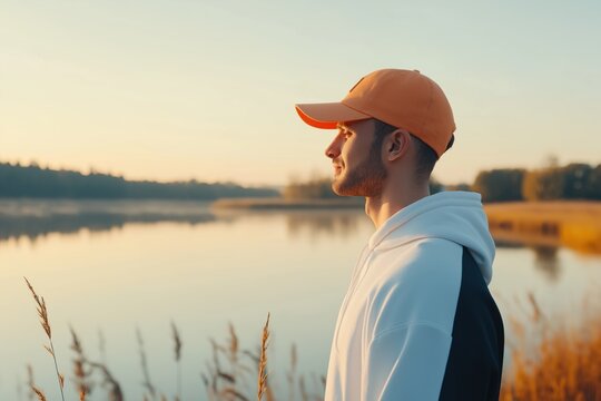 A man wearing an orange hat stands on a grassy bank overlooking a lake. The scene is peaceful and serene, with the man looking out over the water
