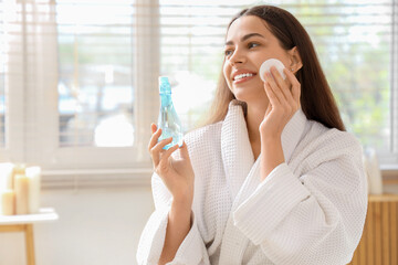 Beautiful young happy woman in bathrobe with cotton pad and bottle of micellar water at home