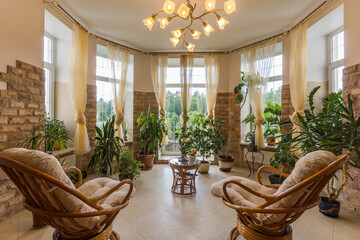 Bay window in a private house with a luxurious interior design. The walls are partially decorated with brick tiles. Wooden furniture, pots with green plants.