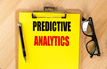 Predictive analytics text on a yellow clipboard with a pen and glasses on a wooden desk