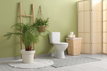 Interior of restroom with toilet bowl, bin, folding screen and plant near green wall