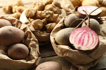Different raw root vegetables in paper bags on wooden table, closeup