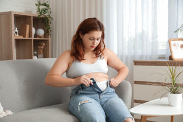 Overweight young sad woman trying to button tight jeans at home. Weight gain concept