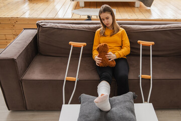 Young girl with broken leg cast sitting on sofa. Sad little kid hugging her teddy bear recovering...