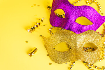Two glittering masks in gold and purple surrounded by festive beads on a bright yellow background,...