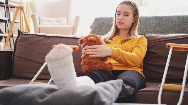 Young girl with broken leg cast sitting on sofa. Sad little kid hugging her teddy bear recovering from broken leg at home. Girl using cast and crutches. Accident injury fracture treatment foot bandage
