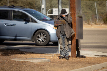 A homeless man wearing a coat on a street corner wiping his face holding a handwritten sign.