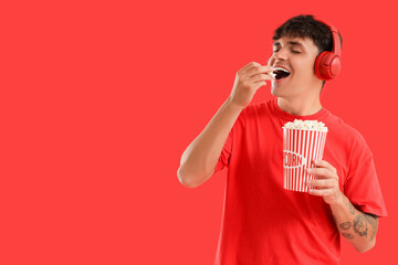 Young man in headphones eating popcorn on red background