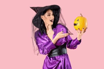 Beautiful young shocked woman dressed as witch for Halloween with pumpkin on pink background