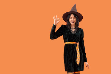 Beautiful young woman dressed as witch for Halloween showing OK gesture on orange background