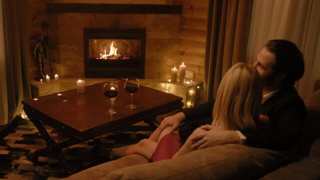 A refined man in a classic suit gently strokes a blonde&rsquo;s knee while she leans closer in a red dress. Wine glasses, glowing candles, and a cozy fireplace create a truly warm atmosphere.
