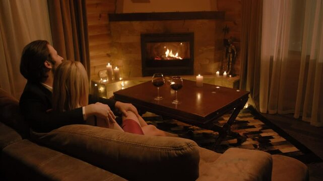 A classic-suited man wraps an arm around a blonde in a red dress, placing his other hand on her knee. Candles glow, wine awaits on the table, and the fireplace casts a warm romantic aura.
