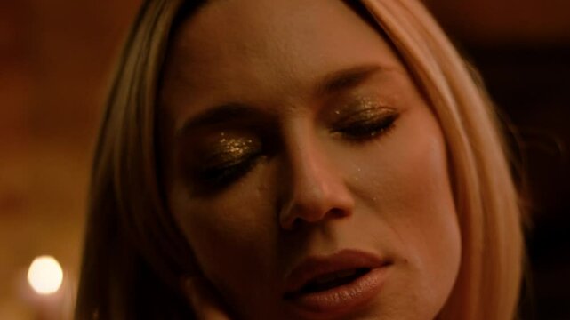 A blonde with loose hair closes her eyes, rolling her head in circles. Soft light illuminates golden eyeshadow, parted lips, and fingertips, while candlelight creates warm intimacy.