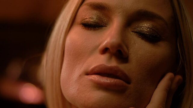 A blonde first gazes forward, then closes her eyes with smooth head circles. Soft light reveals golden eyeshadow, parted lips, and gentle fingertips amid warm candlelight and garlands.