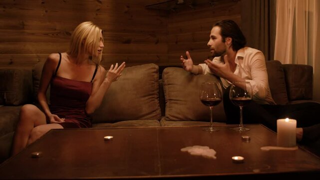 A blonde in a burgundy dress and a brunette man in an open white shirt argue on a beige sofa. She leans forward, gesturing sharply, while he raises a defensive hand near wine and candles.