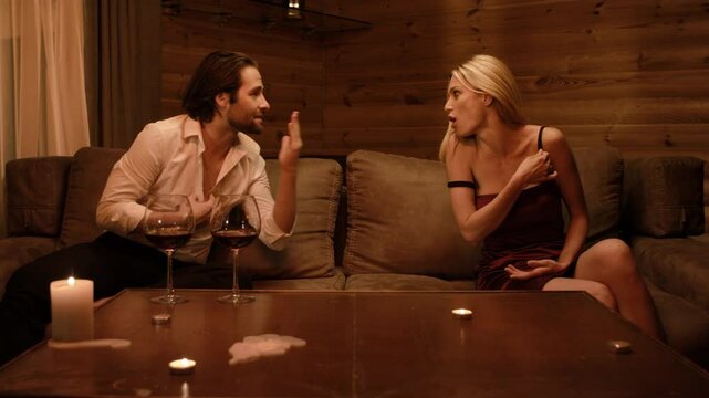 A blonde in a burgundy dress and a stubbled brunette man in an open shirt argue heatedly on a beige sofa. Tension melts into sudden laughter as she shifts from anger to playful teasing.