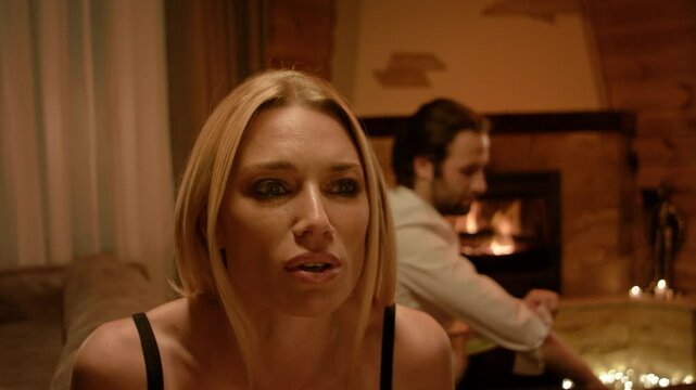 A blonde speaks in a calm yet expressive tone, hands moving gently. A brunette man in a white shirt stands behind, adjusting his sleeves as a fireplace and soft candlelight enhance the tranquil mood.