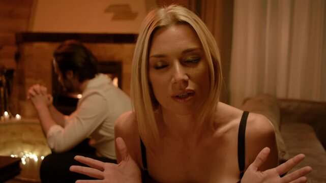 A blonde addresses someone with a calm, measured tone, hands moving smoothly. Behind her, a brunette man in a white shirt rubs his hands quietly. A fireplace and candle glow soften the scene.