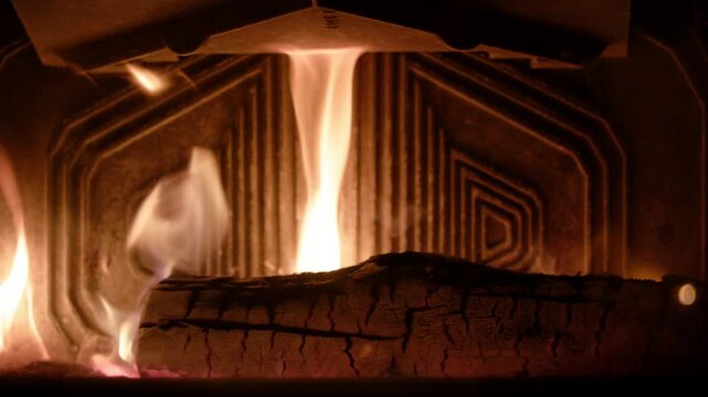 Flames stretch upward in shifting tones from pale white to deep orange, licking a cracked log glowing with red embers. Intricate metal paneling reflects the blaze, intensifying the cozy ambiance.