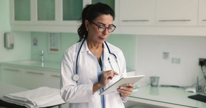 Smiling aged female therapist using tablet working online in medical office, noting important information, manage appointments, provide telemedicine consultations to clinic patients, use e-health apps