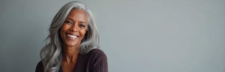 Fototapeta premium Headshot of beautiful mature black woman. Smiling female with glowing skin and long silver hair radiating confidence, natural beauty in neutral setting for skincare, haircare industry.