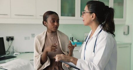 Young African woman patient share health concerns, talk to mature European female general practitioner holding clipboard writing complaint, symptoms, discussing potential diagnoses, tests or procedure