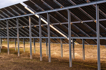 Rows of solar panels on metal frames in a dry area with sparse grass. Shadows suggest late...