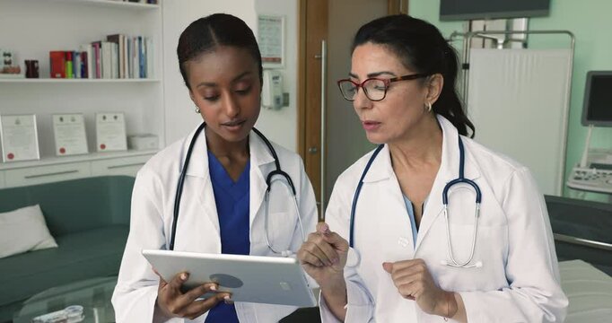 Two multiethnic young and mature women doctors look at tablet screen, discuss diagnostic imaging, review patient medical records, analyzing lab results, considering treatment options, planning surgery
