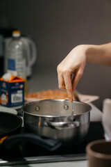 A close-up shot of a hand stirring food in a stainless steel pot on a stovetop, with a blurred background of kitchen ingredients and utensils, capturing a cozy home cooking moment