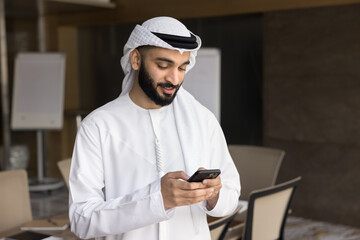 Young 25s businessman dressed in traditional Arabian kandura and ghutra using smartphone, read message, check emails, reviewing information, responding, lead professional communication or making call