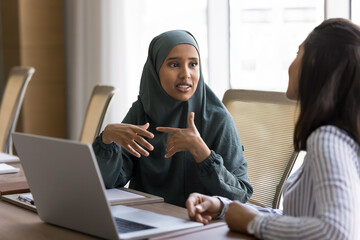 Arabian woman dressed in hijab discuss project updates with Latina colleague, delegating tasks and...