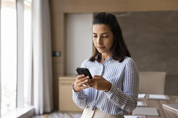 Young 30s Latina businesswoman using smartphone in office, chatting online, share messages, solve...