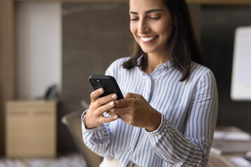 Close up smiling Brazilian businesswoman using smartphone in modern office, engaged in professional tasks, check emails, respond to messages, set reminders, share file or documents, make business call