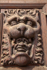 Close-up shot of a weathered, carved wooden panel featuring a grotesque face with an open mouth and bared teeth, serving as an ornamental detail on a piece of furniture or architecture.